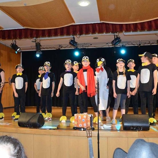 Eine Gruppe von Kindern in Pinguinkostümen steht auf der Bühne, mit Scheinwerfern und Lautsprechern im Hintergrund. Einige tragen Hüte, Schals und Schuhe, während einer einen Koffer hält.