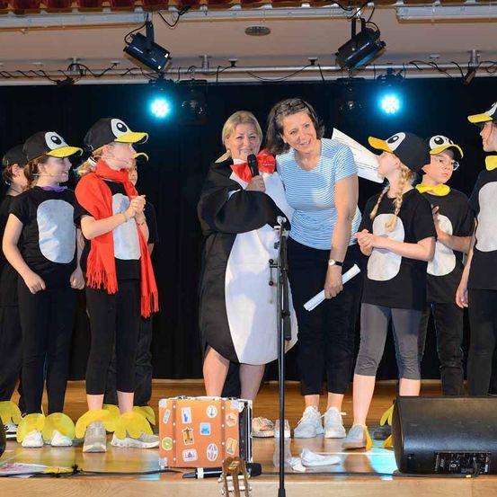 Eine Gruppe von Kindern in Pinguinkostümen steht mit zwei Erwachsenen auf der Bühne, einer hält ein Mikrofon. Die Bühne ist mit blauen und weißen Lichtern beleuchtet.