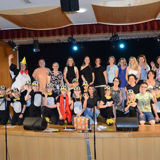 Eine Gruppe von Erwachsenen und Kindern, die in Pinguinkostümen gekleidet sind, posiert für ein Foto auf einer Bühne. Es gibt Musikinstrumente auf der Bühne, und einige der Kinder tragen Brillen.