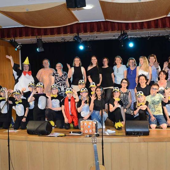 Eine Gruppe von Erwachsenen und Kindern steht auf der Bühne, einige in Pinguinkostümen, andere in Freizeitkleidung. Sie lächeln und posieren für ein Foto.