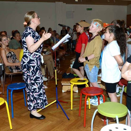 Eine Frau führt eine Gruppe von Kindern in einem Raum mit bunten Stühlen und einem Mikrofonstativ. Eine Frau im Publikum sitzt auf einem Stuhl.