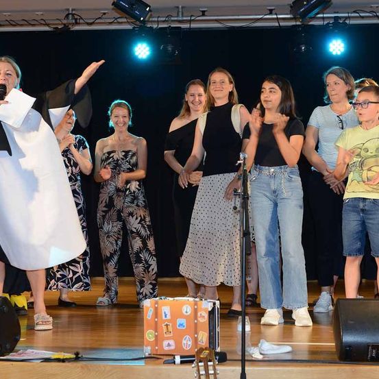 Eine Gruppe von Menschen auf der Bühne, eine hält ein Mikrofon und andere klatschen. Einige tragen lässige Kleidung, und ein Koffer mit Aufklebern steht auf der Bühne.