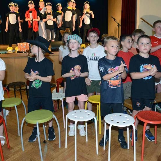 Kinder stehen auf Hockern in einem Klassenzimmer, während andere in Kostümen auf einer Bühne im Hintergrund stehen. Sie scheinen an einer Schulveranstaltung teilzunehmen.