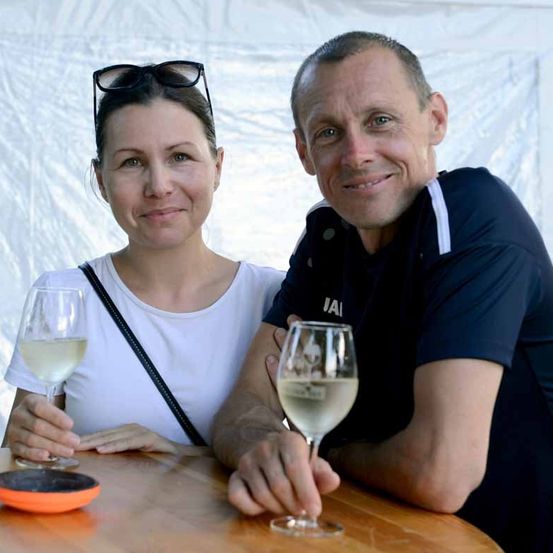 Ein Mann und eine Frau sitzen lächelnd an einem Tisch, jeder hält ein Glas Weißwein. Der Mann trägt ein dunkles Shirt mit weißen Akzenten, und die Frau trägt ein weißes Shirt. Sie sitzen eng beieinander, mit einem weißen Hintergrund hinter ihnen.