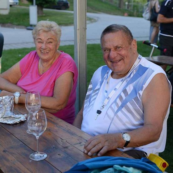 Ein älteres Paar sitzt mit Wein glasses am Tisch und lächelt. Dahinter ist eine Rasenfläche mit einem Weg. Eine weitere Person steht auf dem Weg.