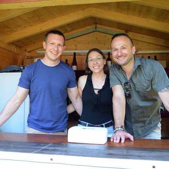 Drei Personen, zwei Männer und eine Frau, lächeln für ein Foto in einem hölzernen Raum. Sie stehen hinter einem Holztresen, mit Flaschen im Hintergrund.