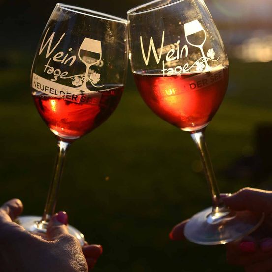 Zwei Hände stoßen mit Weingläsern an, die mit rotem Wein gefüllt sind. Jedes Glas ist mit den Worten 'Wein tage' und einem Weingläser-Symbol graviert. Der Hintergrund ist unscharf.