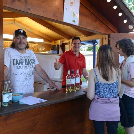 Ein Mann mit einer New York Yankees-Kappe lächelt an einer Bar, an der drei Flaschen Wein ausgestellt sind. Ein Mann in einem roten Hemd gibt einen Daumen hoch. Zwei Frauen stehen vor der Bar.