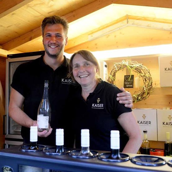 Zwei Personen lächeln in einem Raum mit Holzwänden und -decke, stehen hinter einem Tisch mit Flaschen. Der Mann hält eine Flasche, und die Frau hat ihren Arm um ihn gelegt. Dahinter sind ein Kranz und Kisten mit Kaiser zu sehen.