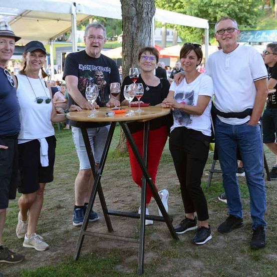Eine Gruppe Erwachsener posiert für ein Foto in einem Park mit einem Tisch im Vordergrund. Zwei Frauen und zwei Männer stehen neben einer Frau mit Brille. Es gibt Weingläser auf dem Tisch.