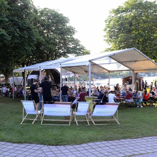 Eine große Versammlung unter einem Zelt in einem Park in der Nähe eines Flusses, mit Menschen, die an Tischen sitzen, einige stehen, und einer Bühne im Hintergrund.