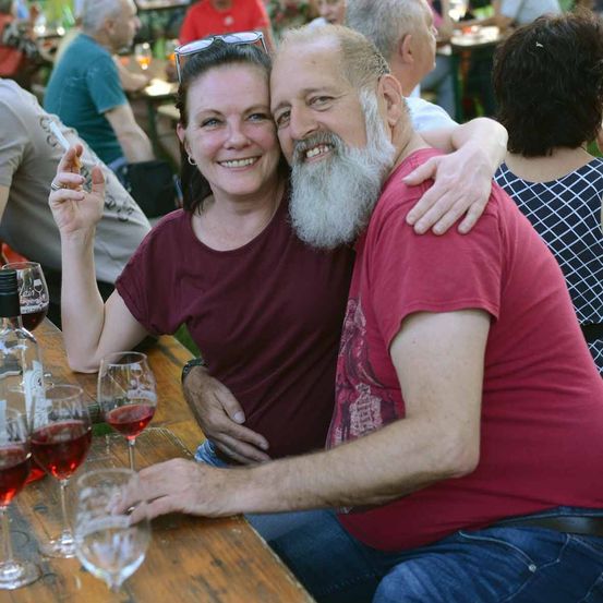 Ein Mann und eine Frau lächeln und posieren für ein Foto in einem Outdoor-Restaurant. Der Mann hat einen Bart und die Frau trägt ein weinrotes Oberteil. Sie sind von anderen Menschen umgeben, von denen einige Wein trinken.