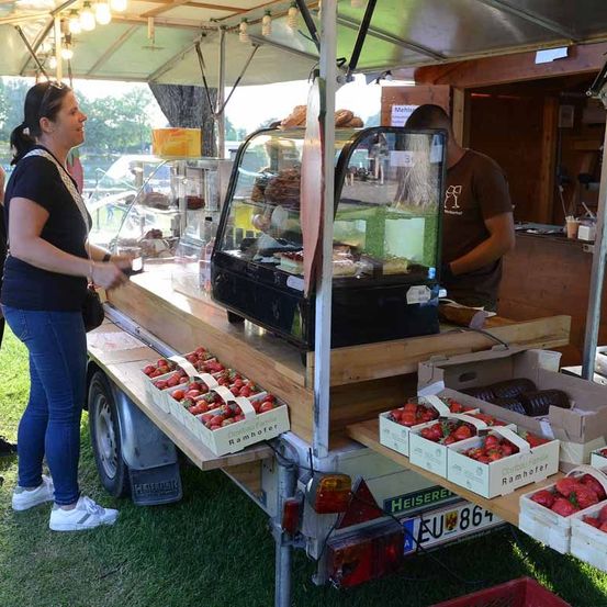 Eine Frau steht vor einem Essenswagen mit Erdbeeren und anderen Früchten. Ein Mann ist im Wagen.