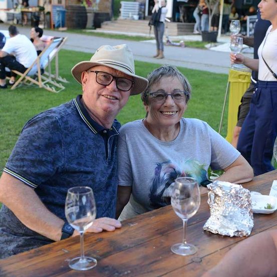 Ein Mann und eine Frau lächeln in die Kamera, während sie an einem Tisch mit Weingläsern und Essen sitzen. Sie sind in einem Park, in dem Leute auf Stühlen sitzen und im Hintergrund spazieren gehen.