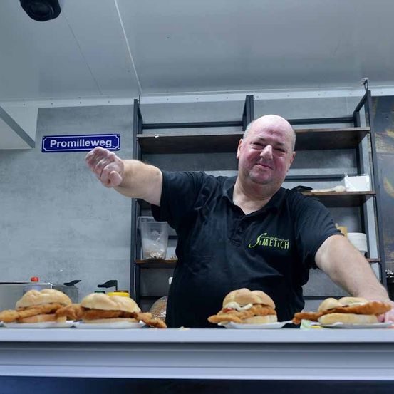 Ein Mann in einem schwarzen Shirt mit Logo steht hinter einem Tresen und präsentiert Essen auf weißen Tellern, mit Regalen und einem Schild mit der Aufschrift 'Promillweg' im Hintergrund.