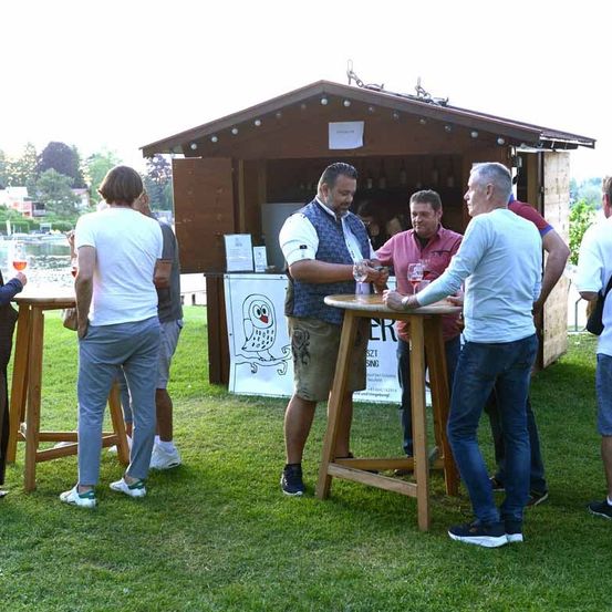 Mehrere Personen stehen um einen kleinen hölzernen Bartresen herum, unterhalten sich und halten Weingläser. Sie sind lässig gekleidet und stehen auf einem grünen Rasen.