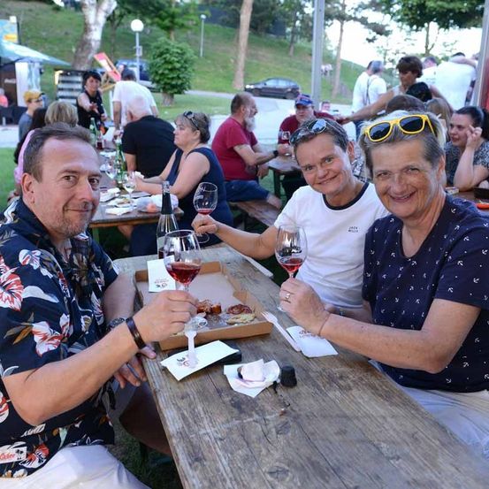 Drei Menschen sitzen an einem Tisch und stoßen mit Weingläsern an. Hinter ihnen sitzen Leute an Tischen und plaudern. Ein Mann trägt einen Ring.