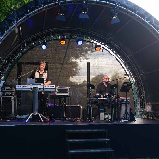 Zwei Musiker spielen auf einer Bühne unter einem Bogen mit Beleuchtung. Einer spielt das Keyboard, der andere die Drums.