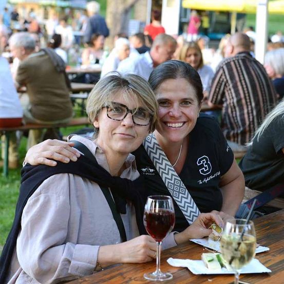 Zwei Frauen sitzen an einem Tisch im Freien, lächeln und posieren für ein Foto. Sie haben Weingläser und Essen auf dem Tisch vor sich. Im Hintergrund sitzen und stehen Leute in einem Außenbereich.