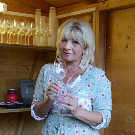 Eine Frau in einem Blumenkleid hält zwei Weingläser in einem Holzraum. Hinter ihr steht ein Regal mit zahlreichen Weingläsern und anderen Gegenständen.