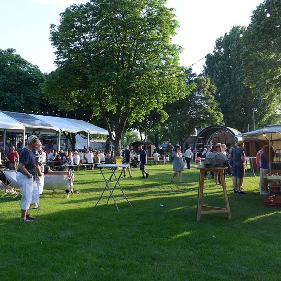 Eine Freiluftveranstaltung mit Menschen, die in einem Park unter Bäumen spazieren und sitzen. Einige stehen in der Nähe von Ständen und Zelten, während andere auf Stühlen sitzen. Der Bereich ist gut beleuchtet.