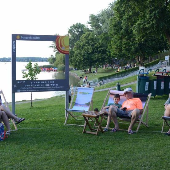 Eine Familie entspannt sich am See. Vater und Sohn sitzen auf Liegestühlen, während die Mutter auf einem Stuhl in der Nähe sitzt. Es gibt ein Schild und einen Fotorahmen in der Nähe.