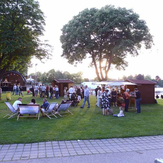Eine Gruppe von Menschen versammelt sich in einem Park für eine Veranstaltung im Freien. Einige sitzen in Klappstühlen, während andere spazieren oder in der Nähe von Ständen und einer Bühne stehen. Bäume und ein See sind im Hintergrund.