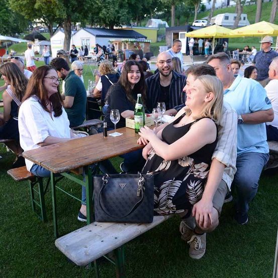 Eine Gruppe von Menschen sitzt auf Bänken um einen Holztisch herum, lächelt und plaudert in einem Außenbereich. Eine Frau hält eine Handtasche. Im Hintergrund sitzen oder stehen weitere Menschen in der Nähe von Essensständen und Zelten.