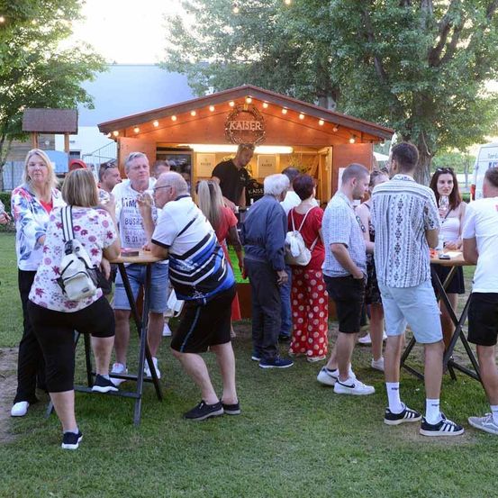 Eine Gruppe von Menschen steht vor einem Kaiser-Kiosk, unter einem Vordach mit Lichtern. Einige halten Weingläser. Ein Mann steht im Inneren des Kiosks. Bäume und ein Gebäude sind im Hintergrund.