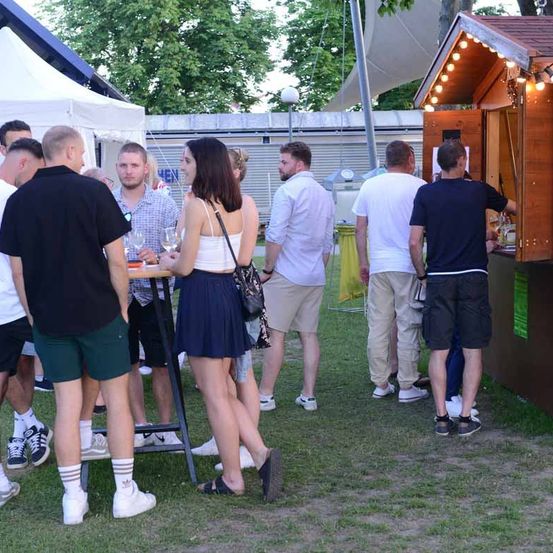 Mehrere Menschen versammeln sich im Freien, möglicherweise bei einer Veranstaltung. Eine Frau in einem blauen Rock steht neben einem Tisch. Hinter ihr unterhält sich ein Mann mit einem anderen. In der Nähe unterhalten sich andere Personen in der Nähe einer Bar. Ein Zelt und Bäume sind im Hintergrund.