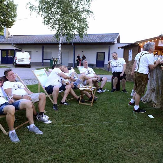 Eine Gruppe von Männern sitzt in Liegestühlen auf einem Rasen vor einem Gebäude, vor dem ein weißer Lieferwagen geparkt ist.
