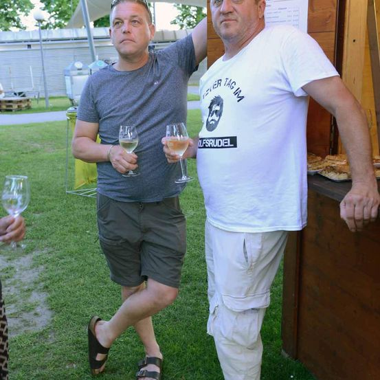Zwei Männer stehen auf einer Rasenfläche. Einer hält Weingläser und trägt Sandalen. Der andere trägt ein weißes T-Shirt mit grafischem Design und legt seinen Arm auf die Schulter des anderen.