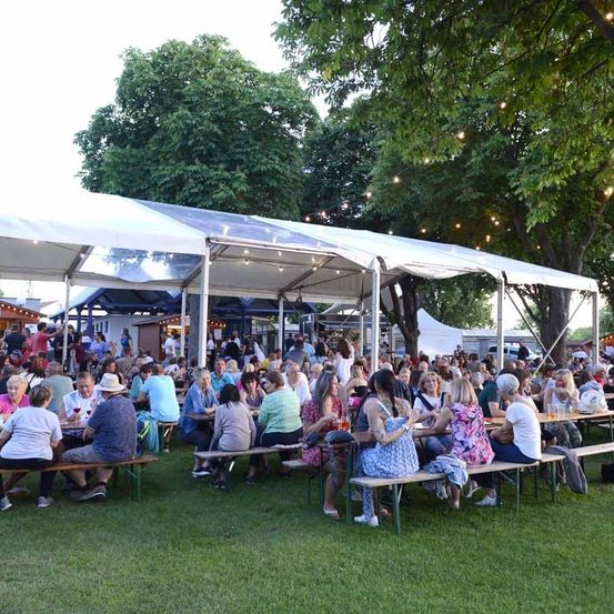 Eine große Menschenmenge sitzt um Tische unter einem Zelt. Sie genießen Getränke und Essen, wahrscheinlich bei einem Festival. Der Bereich ist von Bäumen umgeben und ist im Freien.