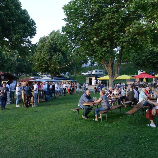 Im Freien sitzen Menschen um Picknicktische unter Schirmen in einem Park. Einige haben Getränke, andere unterhalten sich. Bäume und Zelte sind verstreut. In der Ferne ist ein Gebäude und geparkte Autos zu sehen.