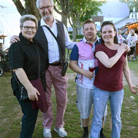 Vier Erwachsene stehen zusammen auf einem Rasen, lächeln und posieren für ein Foto. Der Mann links trägt eine Brille und eine Weste, während die Frau rechts ein Weinglas hält.