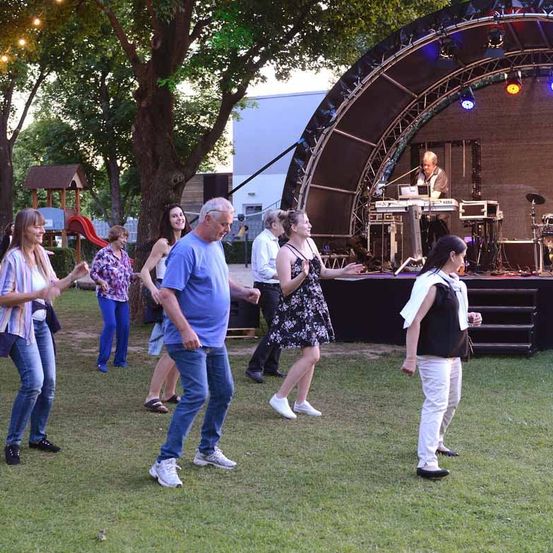 Eine Gruppe von Menschen tanzt auf einem Grasfeld mit einer Bühne dahinter. Die Bühne hat Musikinstrumente und einen Mann, der das Keyboard spielt. Ein Baum und ein Gebäude sind im Hintergrund.
