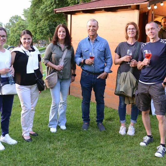 Sechs Menschen stehen draußen auf einer Rasenfläche, lächeln und halten Weingläser in den Händen. Sie sind lässig gekleidet und im Hintergrund steht ein kleines Holzhaus.