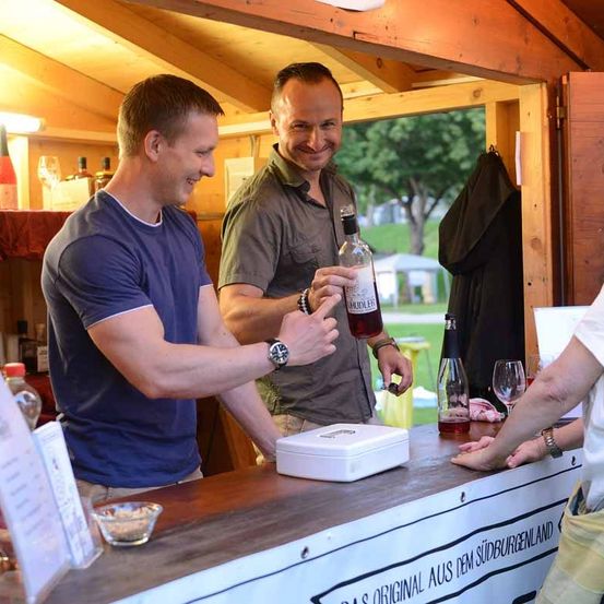 Zwei Männer in einer Bar-Umgebung, einer hält eine Flasche und der andere lächelt, mit einem Schild auf der Theke, das 'Das Original aus dem Sudburgenland' besagt.