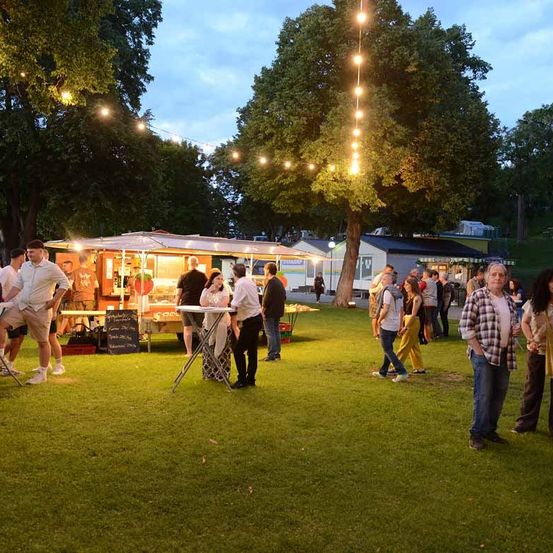 In einem Park versammeln sich mehrere Personen um einen Imbisswagen und einen Tisch mit einer Tafel. Der Bereich ist mit Lichterketten geschmückt.