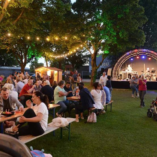 Eine Gruppe von Menschen sitzt auf Bänken in einem Außenbereich. Einige von ihnen trinken Wein. Vor einer Bühne stehen mehrere Menschen. Die Bühne hat einige Musikinstrumente.