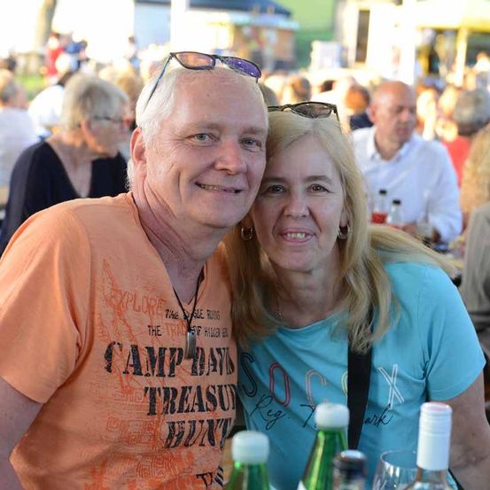 Ein Paar in einem Außenbereich mit vielen Menschen. Der Mann trägt ein orangefarbenes Shirt mit 'Camp David' Schriftzug und die Frau ein blaues Shirt. Sie lächeln. Dahinter stehen Flaschen, Weingläser und ein unscharfer Hintergrund.