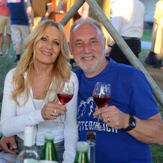 Ein Mann und eine Frau lächeln und halten Weinpokale bei einer Outdoor-Veranstaltung. Sie sitzen vor Flaschen und einer Metallstruktur. Im Hintergrund stehen Menschen.