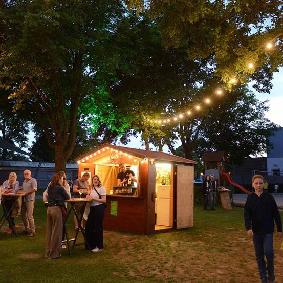 Eine Außenbar ist nachts beleuchtet, mit mehreren Menschen, die herumstehen. Ein Junge geht an der Bar vorbei, wo ein Mann Getränke serviert. Bäume und ein Spielplatz sind im Hintergrund.