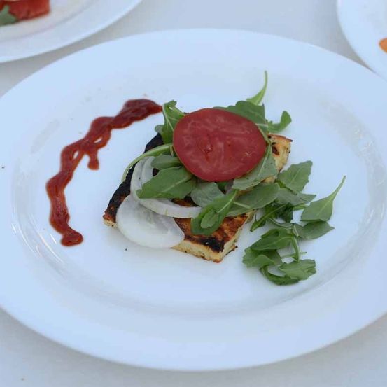 Ein gegrilltes Brotquadrat mit Tomatenscheibe, Rucola und Zwiebel garniert mit roter Sauce auf einem weißen Teller.