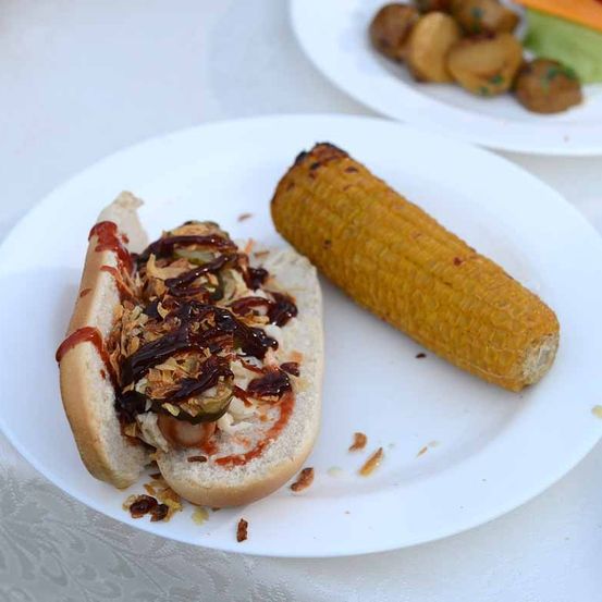 Ein Teller mit einem halb gegessenen Hot Dog, der mit einer scharfen Sauce, Maiskolben und anderen Toppings belegt ist, und ein Teller mit gebratenen Kartoffeln.