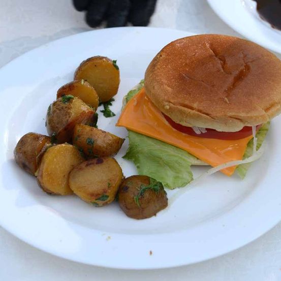 Ein weißer Teller hat einen Cheeseburger mit Salat, Tomate und Käse oben und eine Beilage aus gebratenen Kartoffeln mit Kräutern.