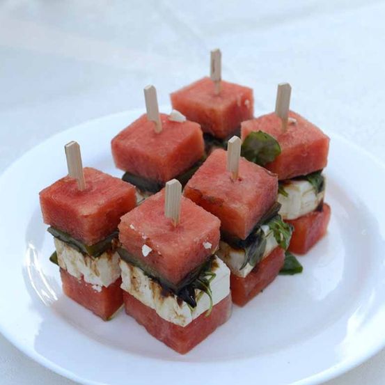 Ein Teller mit sechs Wassermelonenwürfeln, jeweils mit Käse, Grünzeug und einem Stiel belegt. Sie sehen frisch und appetitanregend aus.