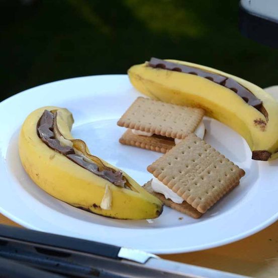 Ein weißer Teller hat einen Bananensplit mit Schokolade, zwei Graham-Cracker mit Marshmallows und einen Spatel.