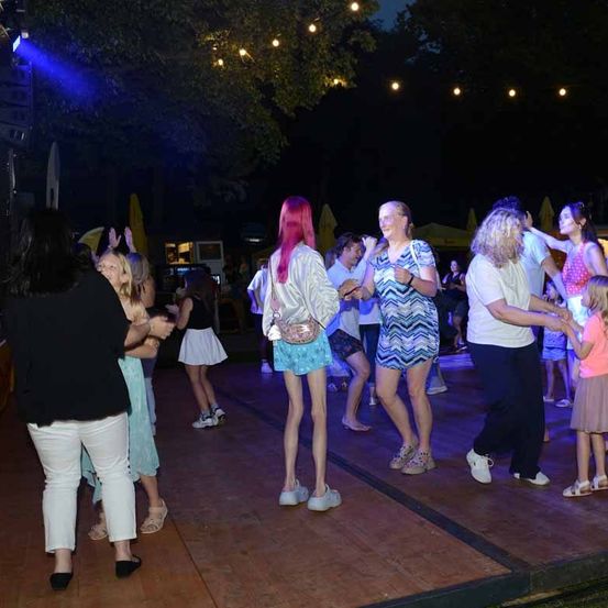 Eine Gruppe von Frauen und Kindern tanzt bei Nacht auf einem Holzboden unter Lichterketten.