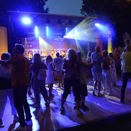Eine Gruppe von Menschen tanzt bei Nacht auf einer Bühne unter blauen und weißen Lichtern. Eine Band spielt auf der Bühne.